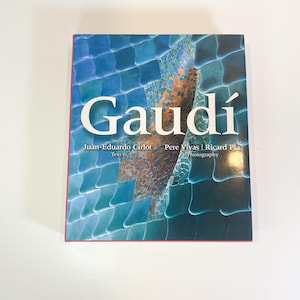 May include: A hardcover book titled "Gaudí" with a cover featuring a mosaic design in shades of blue, teal, and brown. The title is in large white letters. The author's names are also visible.