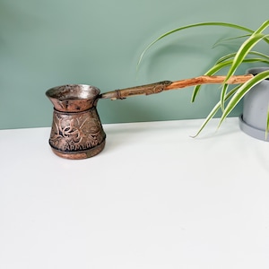 May include: A vintage copper coffee pot with a long wooden handle. The pot has ornate floral and leaf detailing with the word "Armenia" inscribed. The pot is a warm copper color and sits on a white surface.