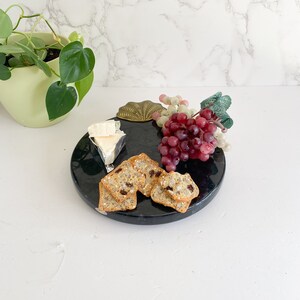 May include: A round, black marble serving board with crackers, cheese, and grapes. A decorative brass shell is on the board. A green plant in a light green pot is in the background.
