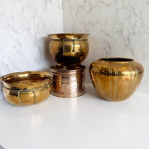 May include: Four antique brass planters of varying sizes and shapes. The planters have a hammered texture and a warm, golden hue. One planter has a rope detail. The planters are arranged on a white surface against a marble background.