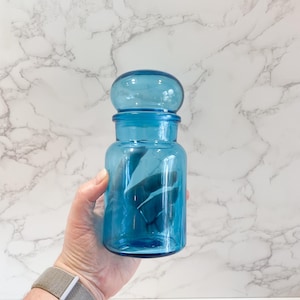 May include: A blue glass jar with a rounded lid. The jar is clear and has a textured surface.
