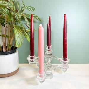 May include: A clear glass candelabra holds five candles. Four are tall and burgundy, and one is pink. The candelabra is on a white surface with a green wall in the background. A potted plant is to the left.