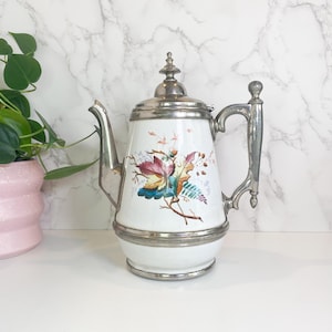 May include: White ceramic teapot with a silver metal handle, spout, and lid. The teapot features a colorful floral design with red, yellow, blue, and green leaves. The teapot has a bulbous base and a decorative finial on the lid.