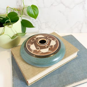 May include: A ceramic, teal-colored vessel with a brown center featuring white animal figures. The vessel has a central hole and sits atop two books. A green potted plant is visible in the background.