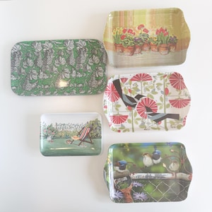 May include: A collection of five decorative trays with various designs. One tray features a green grape pattern, another showcases potted flowers, and a third displays birds. The remaining trays depict a garden scene with a chair and a bird scene.