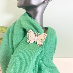 May include: A silver-toned butterfly brooch with intricate wing detailing. The brooch is pinned to a vibrant green fabric, showcasing its delicate design. The butterfly's wings have a textured appearance, and the brooch is displayed on a mannequin.