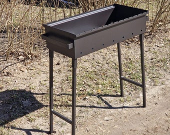 May include: A dark gray metal barbecue grill with a rectangular firebox and a lower shelf. The grill has four legs and is designed for outdoor cooking. The firebox has small holes along the sides for ventilation.