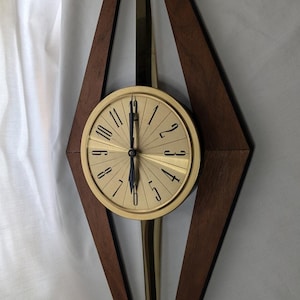 Puede incluir: Un reloj de pared con una esfera circular de tono dorado con números y manecillas negras, montado dentro de un marco vertical en forma de diamante hecho de madera marrón oscura. Un eje metálico dorado se extiende verticalmente por el centro.
