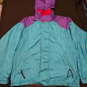 May include: A teal and purple windbreaker jacket featuring a detachable hood, a high collar with a bright pink interior lining, a front zipper closure with a snap-button placket, two zippered side pockets, and a small black brand tag on the chest.