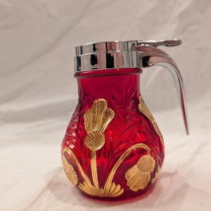Op de afbeelding: Een siroopdispenser van robijnrood glas met een metalen chromen deksel en een hendel voor duimbediening. De glazen behuizing is versierd met in reliëf aangebrachte goudkleurige distelbloemmotieven die rond de buitenkant van de houder lopen.