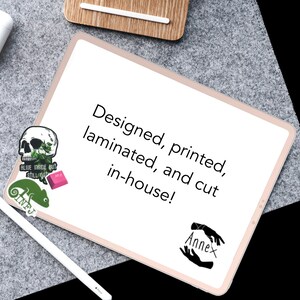 May include: A white tablet with a pink border, displaying the text "Designed, printed, laminated, and cut in-house!" with a black and white hand illustration with the word "Annex" below. There are three stickers on the tablet: a skull with green leaves, a pink rectangle, and a green chameleon.
