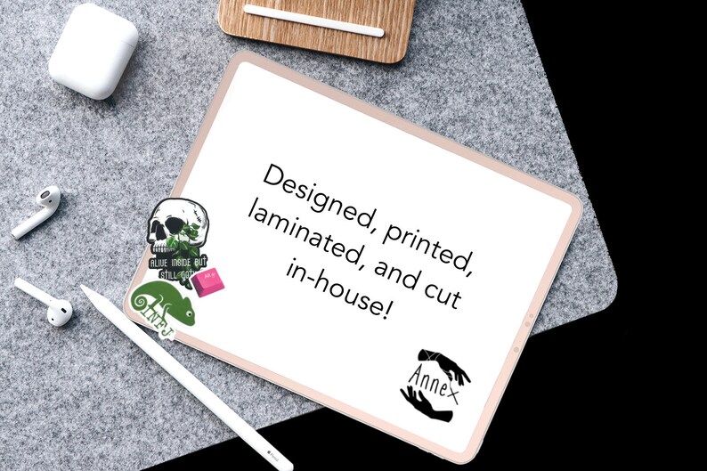May include: A white tablet with a pink border, displaying the text "Designed, printed, laminated, and cut in-house!" with a black and white hand illustration with the word "Annex" below. There are three stickers on the tablet: a skull with green leaves, a pink rectangle, and a green chameleon.