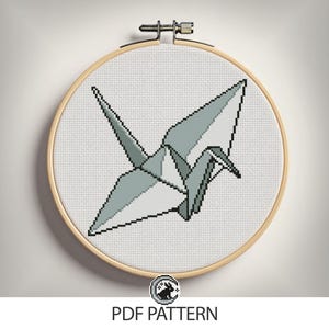 以下が含まれることがあります： 木製の刺繍枠に収められた、灰色の折り紙の鶴のクロスステッチパターン。 鶴は白い布の背景に配置されています。 画像の下部には「PDF PATTERN」というテキストがあります。