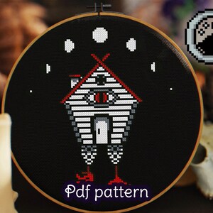 May include: A black and white cross-stitch pattern of a house with an eye on the front, standing on two legs with red shoes. The house is surrounded by five white moons on a black background. The text "Pdf pattern" is written below the house.