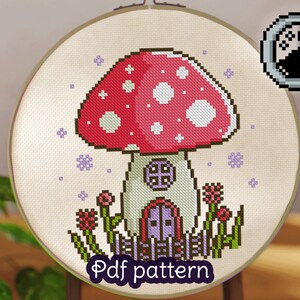 May include: Cross stitch pattern of a mushroom house with a red and white mushroom cap, a white house with a purple door, and green grass and flowers. The pattern is on a white background with purple snowflakes. The text "Pdf pattern" is at the bottom of the image.