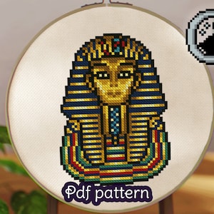 May include: A cross-stitch pattern of a gold pharaoh's mask with blue and red accents. The pattern is displayed on a white canvas in a round embroidery hoop. The text "Pdf pattern" is written below the pattern.