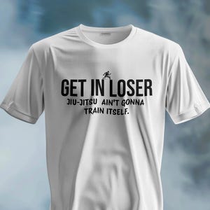 May include: White t-shirt featuring the text "GET IN LOSER" in black, bold font. Underneath, it reads "JIU-JITSU AIN'T GONNA TRAIN ITSELF." A small black silhouette of a person running is above the word "LOSER".