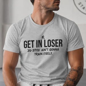 May include: A light grey t-shirt with the words "GET IN LOSER" and "JIU-JITSU AIN'T GONNA TRAIN ITSELF" printed in black. A small graphic of a person is above the word "LOSER". The shirt is made of a soft material.