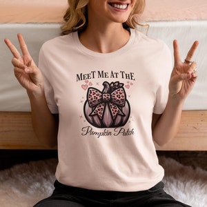 以下が含まれることがあります： ピンクのTシャツに、ピンクと黒のレオパード柄のリボンが付いたカボチャのグラフィックがプリントされています。Tシャツには「Meet me at the Pumpkin Patch」という文字がプリントされています。