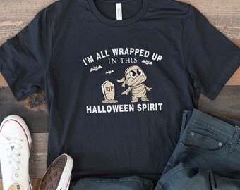Puede incluir: Camiseta azul marino oscuro con el texto "I'M ALL WRAPPED UP IN THIS HALLOWEEN SPIRIT" y un gráfico de momia. La camiseta se combina con vaqueros azules y zapatillas de lona blancas, ideal para un atuendo casual de Halloween.