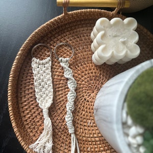 Puede incluir: Dos llaveros de macramé blanco con anillas de plata. Los llaveros están colgados en una bandeja tejida con una vela blanca y una planta en maceta.