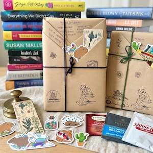 May include: Two books wrapped in brown paper with hand-drawn illustrations and twine, featuring handwritten notes, stickers of a capybara, a cactus, and a bottle of candy, alongside tea bags, a Swiss Miss packet, and a bookmark on a linen surface.