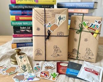 Puede incluir: Dos libros envueltos en papel marrón con ilustraciones dibujadas a mano y cordel, con notas escritas a mano, pegatinas de un capibara, un cactus y una botella de dulces, junto a bolsitas de té, un sobre de Swiss Miss y un marcapáginas.