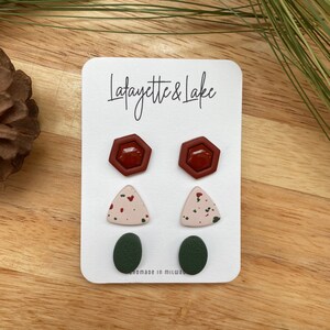 May include: A set of six stud earrings on a white card with the text "Lafayette & Lake" and "Handmade in Milwaukee". The earrings are in three different shapes: hexagon, triangle, and oval. The hexagon earrings are a deep red color, the triangle earrings are a light pink with red and green speckles, and the oval earrings are a dark green.