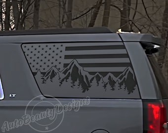 May include: A grey SUV with a black American flag and mountain silhouette decal on the rear window. The decal features a mountain range with pine trees in front of the flag.