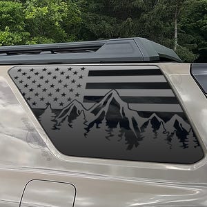 May include: A black graphic of the American flag, mountains, and trees is displayed on a vehicle window. The design features the stars and stripes of the flag, with a mountain range and forest silhouette below.