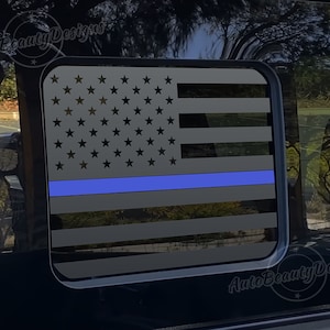 May include: A black and gray American flag with a thin blue line running horizontally across the middle of the flag. The flag is displayed on a car window.
