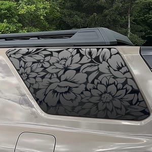 May include: A tan-colored vehicle features a dark gray floral decal applied to the rear side window. The decal displays a repeating pattern of large, stylized flowers and leaves that covers the entire glass surface of the rear quarter window.