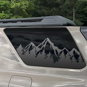 May include: A vehicle window decal featuring a mountain range and forest silhouette. The design is in a dark gray color, with the mountains and trees contrasting against the dark window tint. The decal is applied to the rear window of a vehicle.