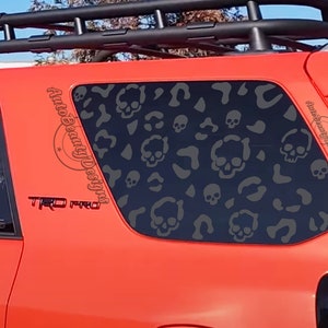 May include: A red SUV with a black roof rack and a skull and crossbones pattern on the rear window. The window decal says "Auto Beauty Designs". The vehicle has a "TRD PRO" emblem on the side.