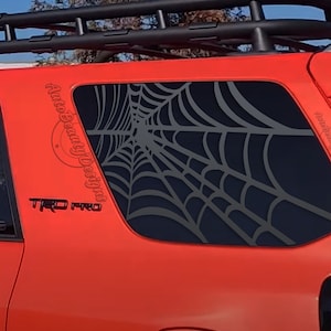 May include: A red car with a black spiderweb decal on the back window. The decal is on the passenger side window and says "Auto Beauty Designs" on the side. The car also has a black "TRD PRO" emblem on the side.