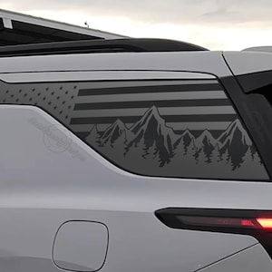 May include: A black and gray graphic decal applied to the rear window of a white SUV. The design features an American flag with a mountain range and forest silhouette. The decal is a decorative accessory for vehicles.