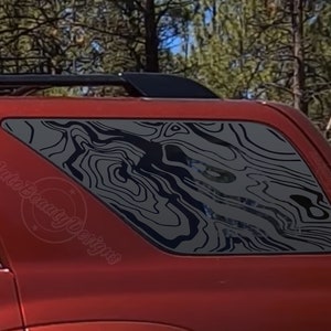 May include: A red SUV with a black roof rack and a rear window with a black and gray topographic pattern decal. The decal has a wavy, abstract design.