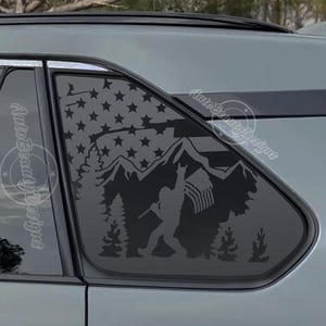 May include: Black decal applied to a car window, featuring a silhouette of a mountain range, American flag, and a figure holding a flag. The design includes trees and stars, creating a patriotic and outdoor theme.