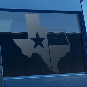 May include: A black and white decal of the state of Texas with a white star in the center. The decal is on a blue car window.