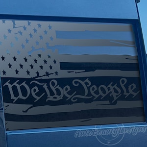 May include: A distressed American flag decal with the words "We the People" in a vintage font. The decal is applied to the rear window of a blue vehicle.