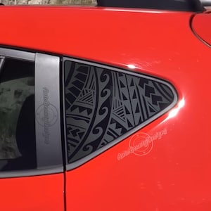May include: A red car with a black tribal design on the rear window. The design is a geometric pattern with a wave-like element. The text "Auto Beauty Designs" is visible on the window.