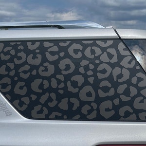 May include: A silver car with a black leopard print decal on the back window. The decal covers the entire window and has a dark gray color.