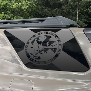 May include: A dark gray vinyl decal applied to the rear side window of a tan vehicle. The decal features the Great Seal of the State of Florida, including the text Great Seal of the State of Florida and In God We Trust, over a cross design.