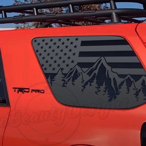 May include: A red SUV with a black roof rack and a black and gray American flag and mountain scene decal on the rear window. The decal features a mountain range with pine trees in front of the flag. The vehicle has a "TRD PRO" emblem on the side.
