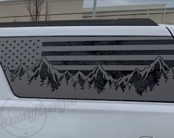 May include: A gray and black American flag decal with mountains and trees in the bottom. The decal is applied to the back window of a white SUV.
