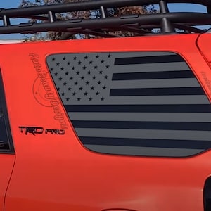 May include: A red Toyota 4Runner with a black roof rack and a black and gray American flag decal on the rear window. The decal has the text "Auto Beauty Designs" on the side. The car has a "TRD Pro" emblem on the side.