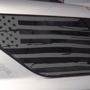 May include: A black and gray distressed American flag decal on the rear window of a silver SUV. The decal is on the back passenger side window. The decal has a distressed look with faded edges and a worn appearance. The decal is a popular choice for car enthusiasts who want to show their patriotism.