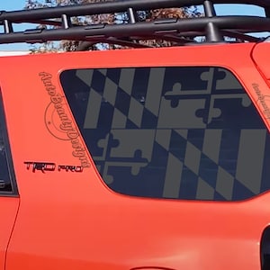 May include: A red SUV with a black roof rack and a black and gold Maryland state flag decal on the rear window. The decal is on the passenger side rear window and has the text "Auto Beauty Designs" on the side. The vehicle also has a black "TRD PRO" emblem on the side.