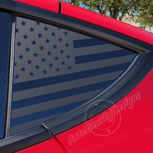 May include: A red car with a black American flag decal on the rear window. The flag is in a gray and blue color scheme. The decal is on the back passenger side window.