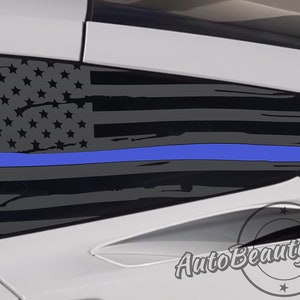May include: A gray and black American flag with a thin blue line across the middle, applied to the rear window of a white car. The flag is distressed and has a faded look. The text "AutoBeautyDesigns" is visible in the lower right corner of the image.